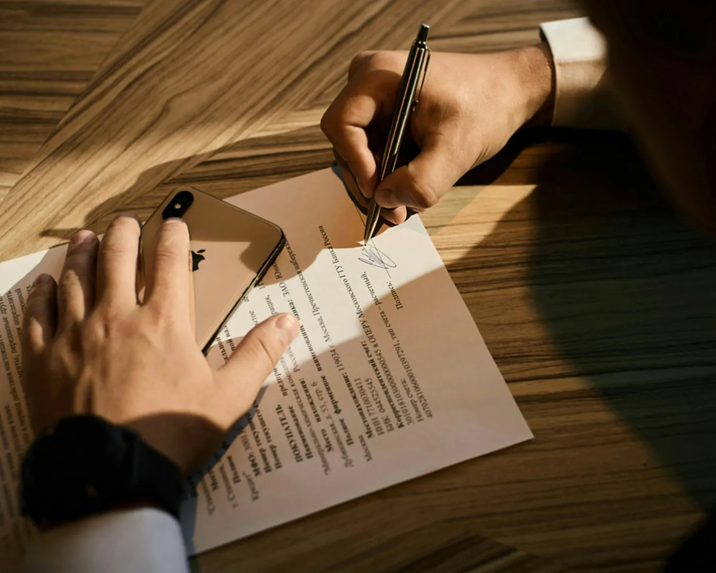 A hand signing a printed document on a wooden desk in late afternoon light.